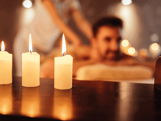 Ein Mann liegt im Hintergrund auf einer Massagebank, im Vordergrund brennen drei weißen Kerzen