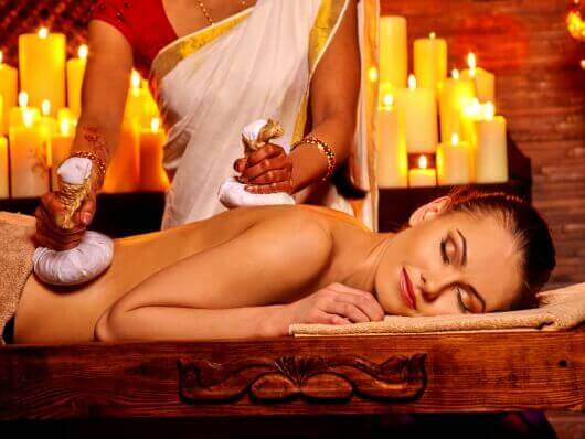 Frau auf einer Massagebank. Brennende Kerzen im Hintergrund. Weitere Frau stehend, gibt eine Kraüterbeutelmassage.